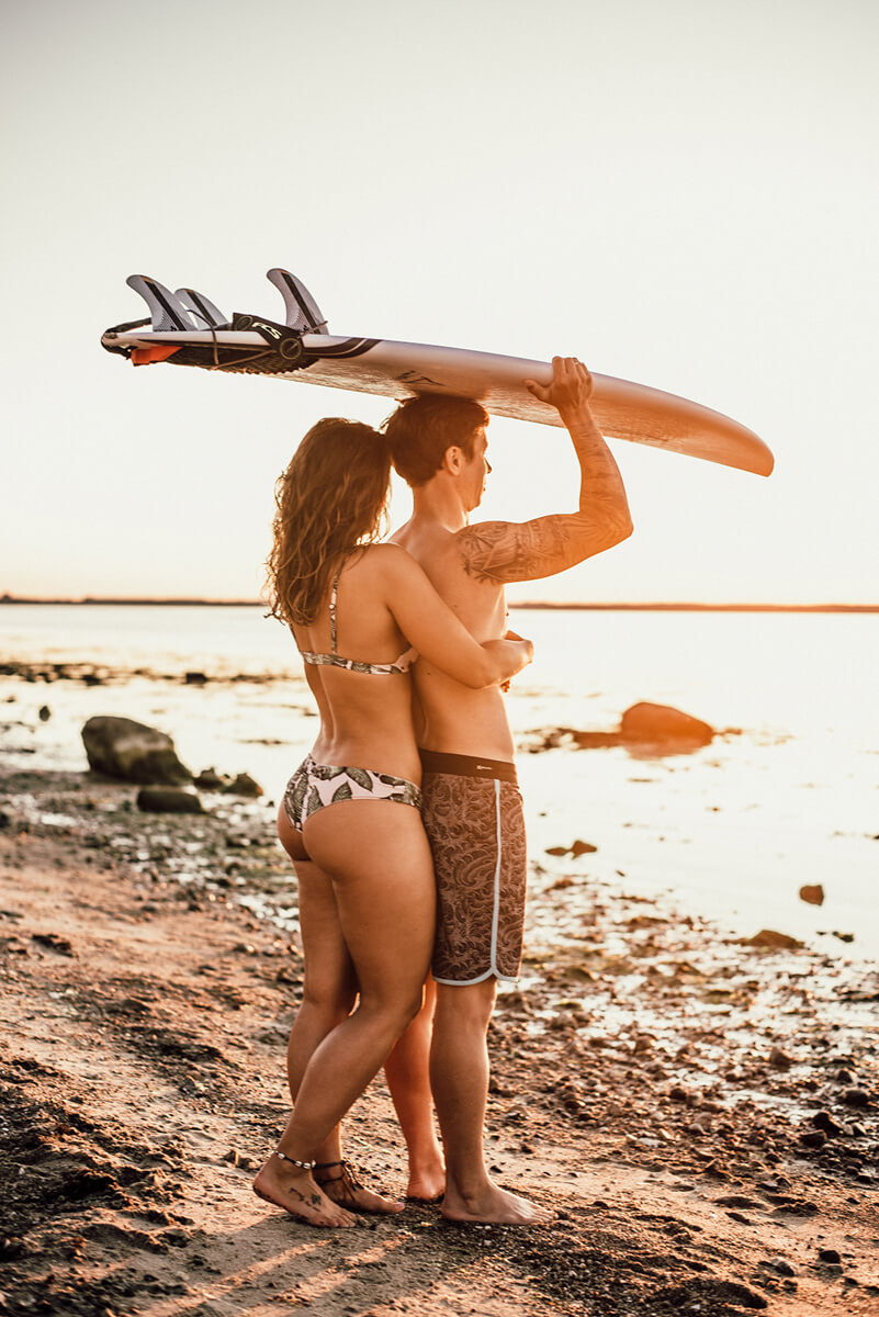 schöne Frau umarmt ihren Partner im Sonnenuntergang von hinten, dieser trägt ein Surfboard auf dem Kopf