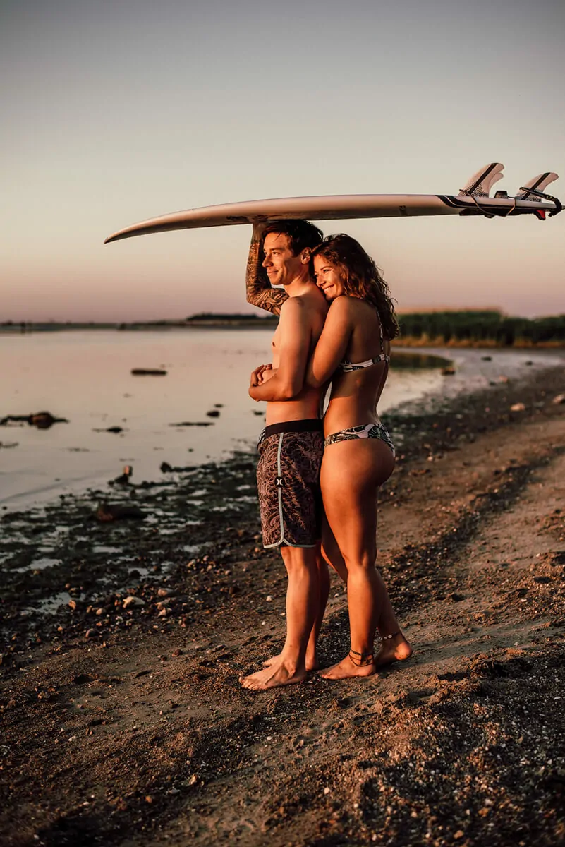 schöne Frau umarmt ihren Partner im Sonnenuntergang von hinten, dieser trägt ein Surfboard auf dem Kopf