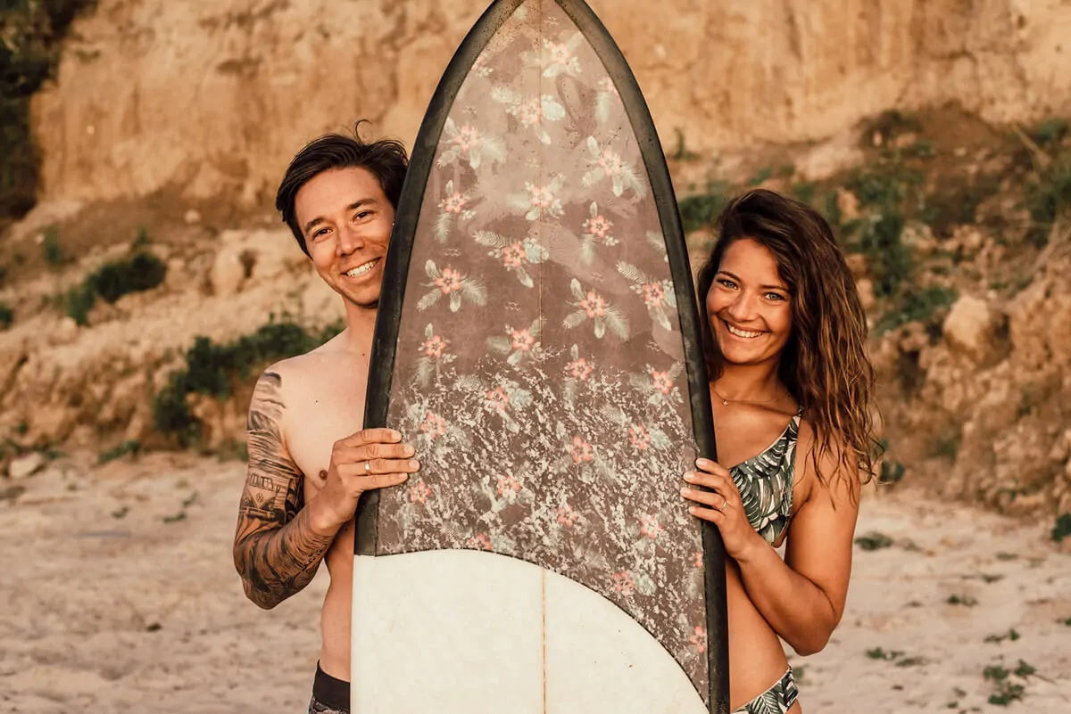 Paar lächelt in die Kamera mit Surfbrett in der Hand